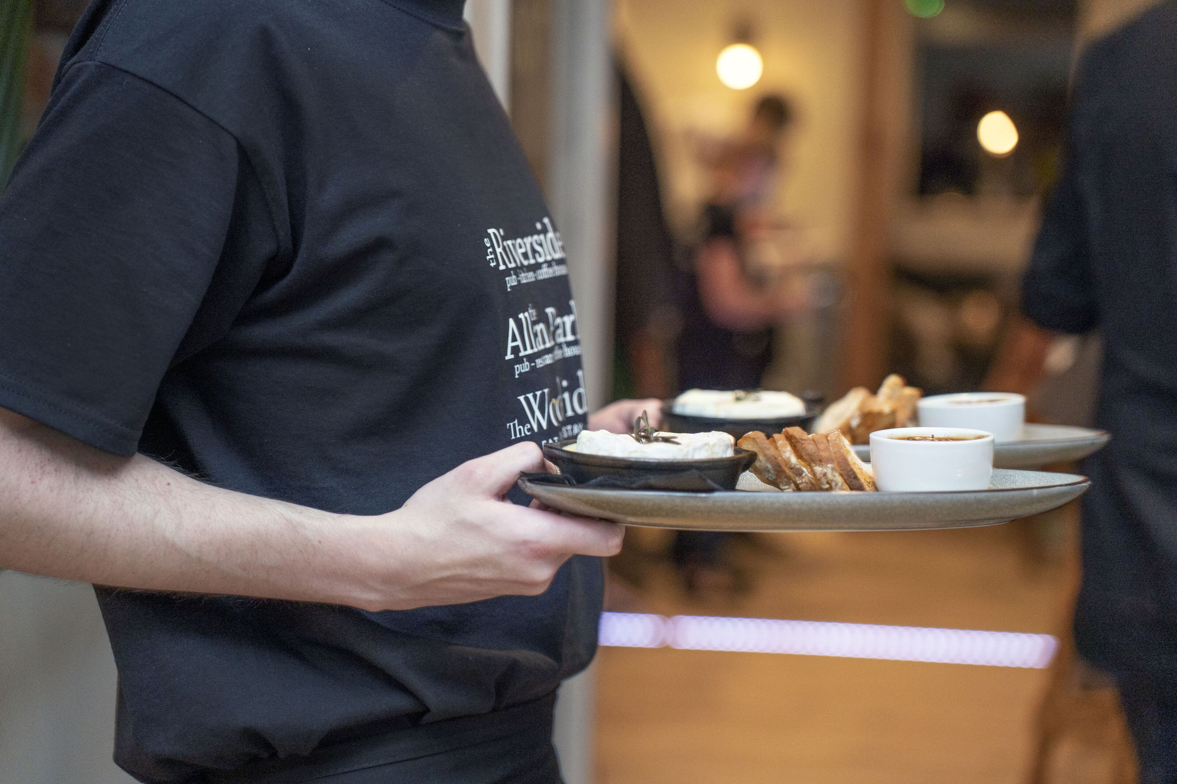 A person holding a tray with food and drinks, wearing a black shirt with text on the back.