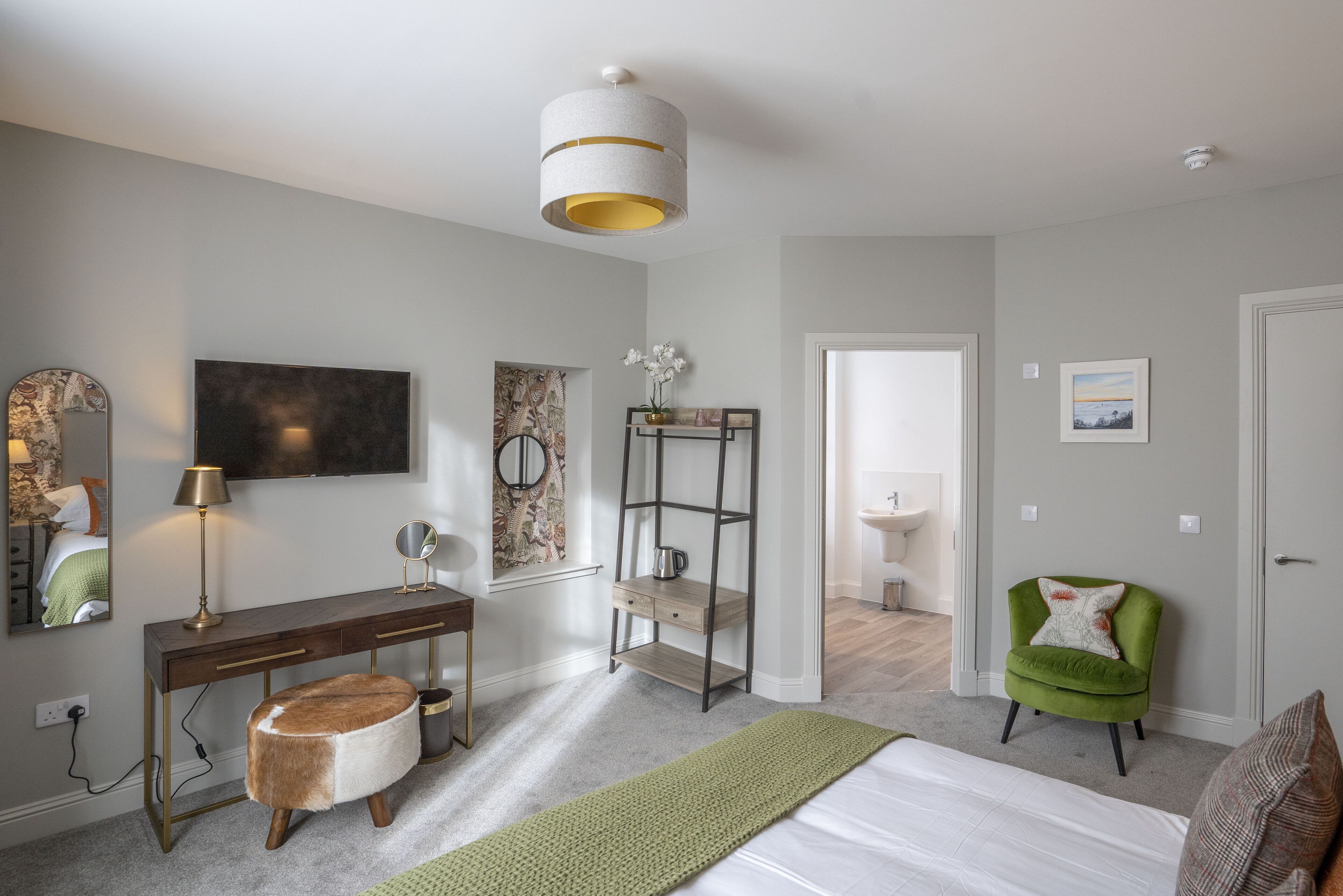 A modern bedroom with a bed, a vanity with a mirror, a green armchair, and a hanging light fixture.