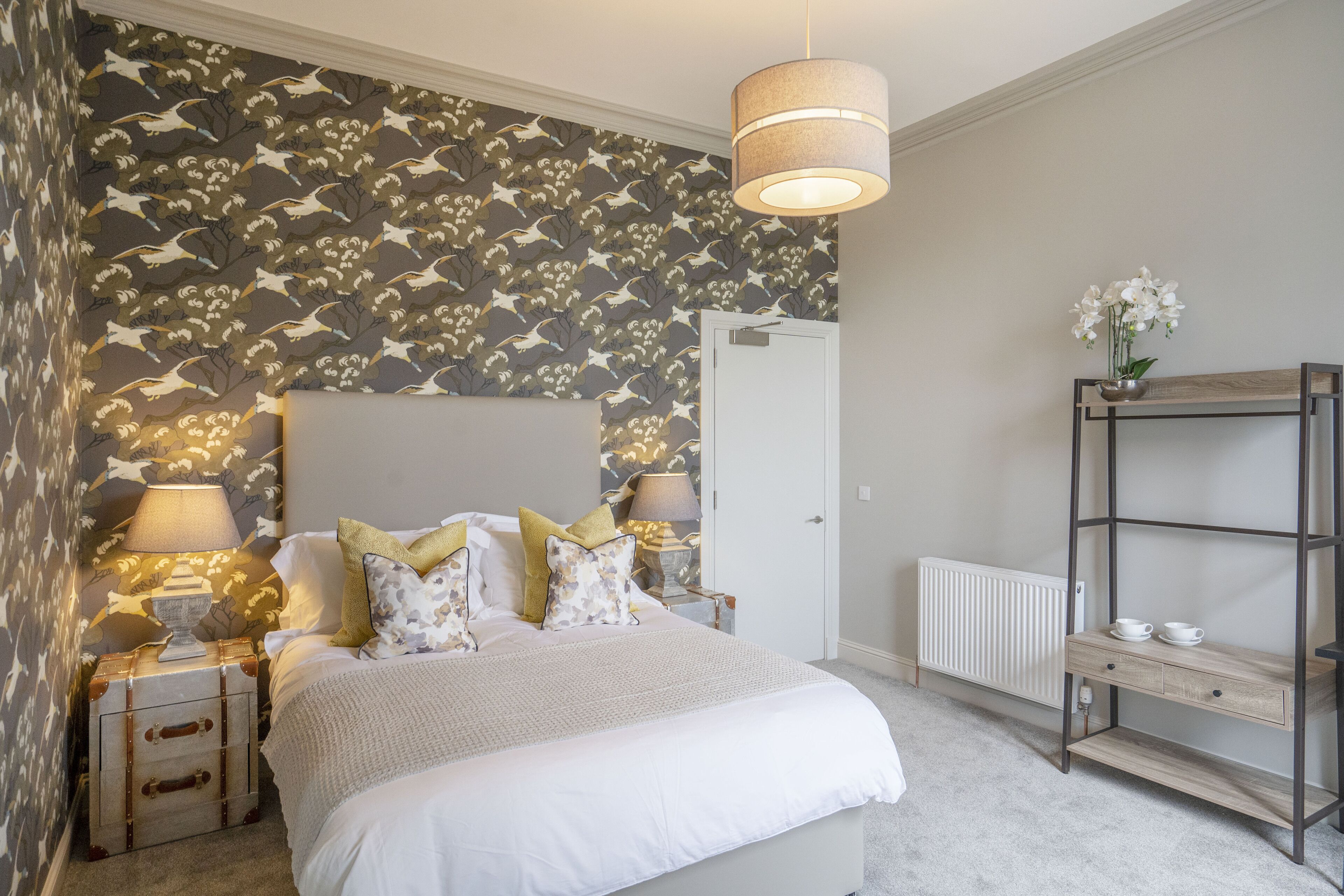 Un dormitorio con papel tapiz estampado, una cama con ropa de cama blanca, dos lámparas de noche, un radiador y una estantería de madera con un jarrón de flores.