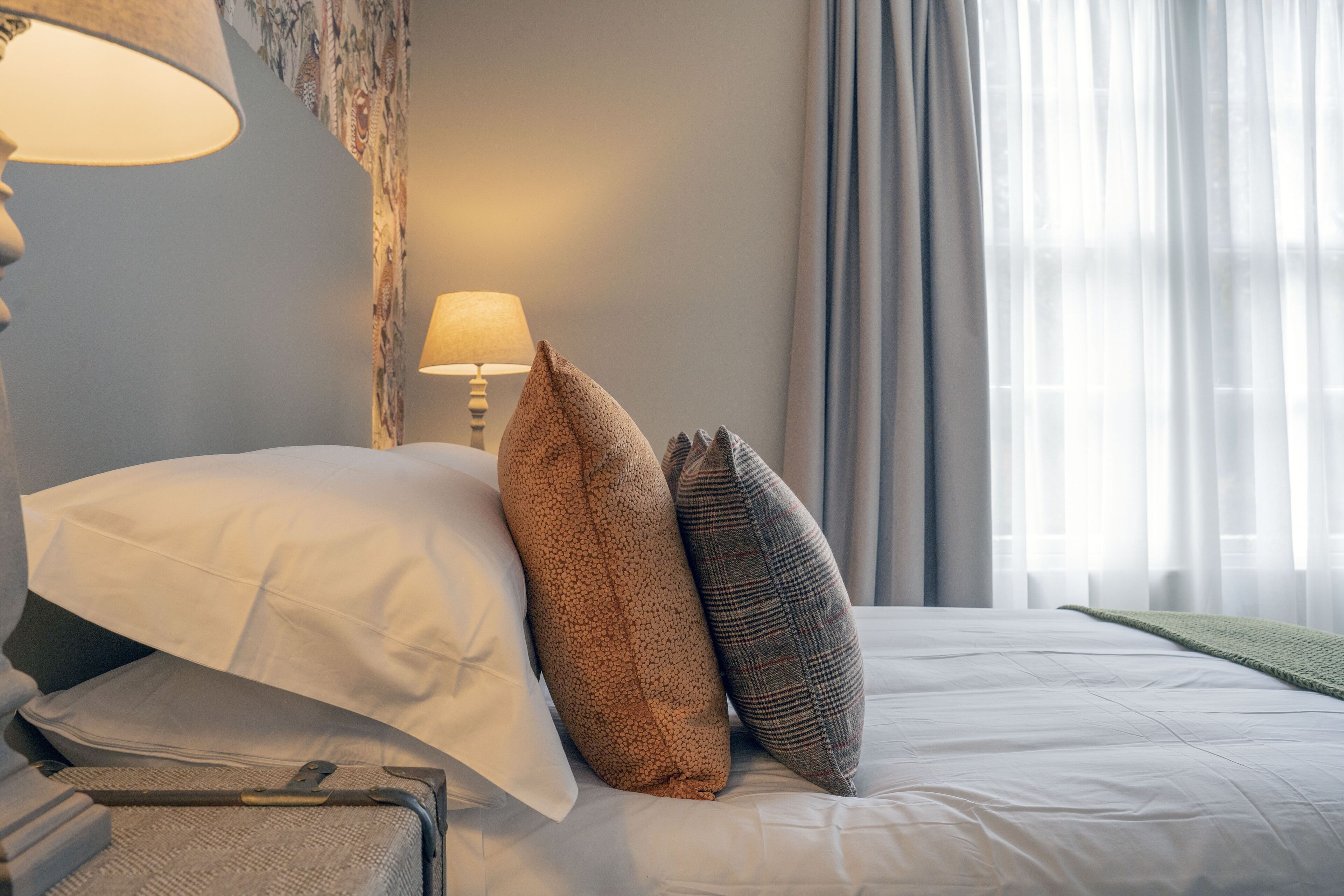 Una cama bien hecha con almohadas y una maleta, una lámpara y una ventana con cortinas.