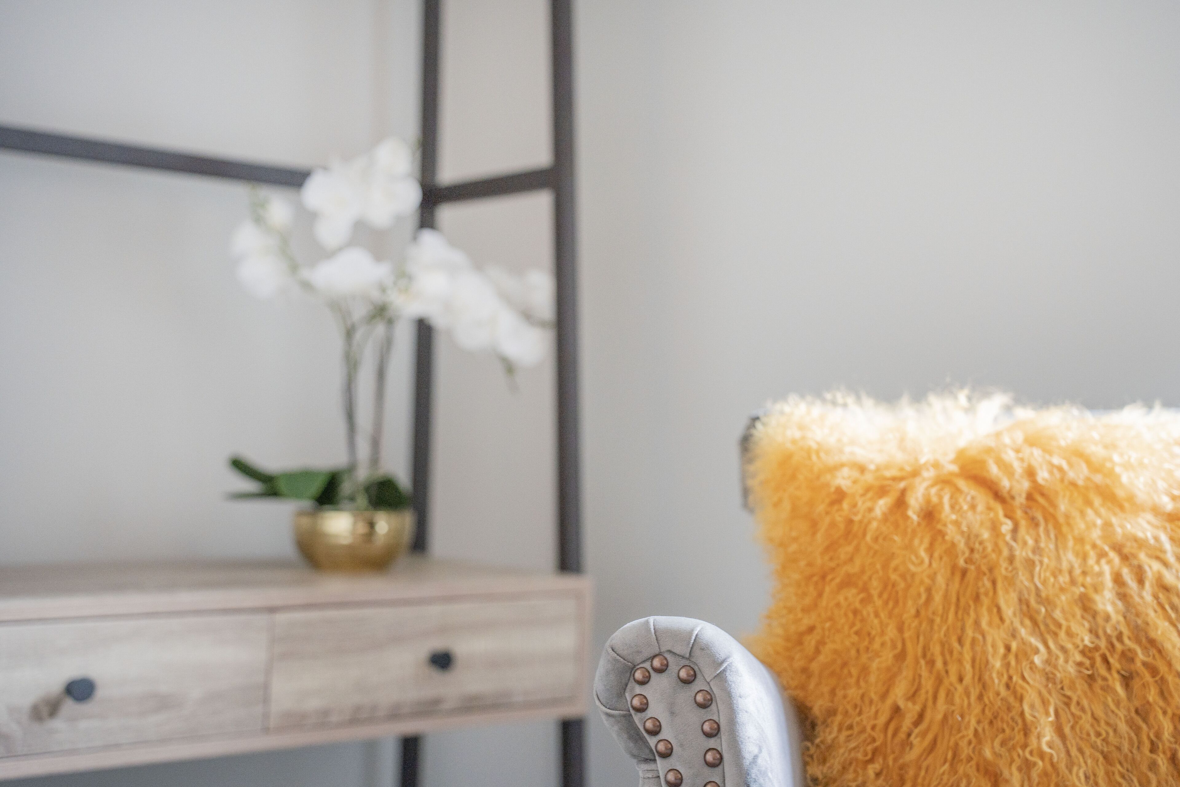 A close-up of a chair with a fluffy cushion, a wooden desk with drawers, and a vase with white flowers.