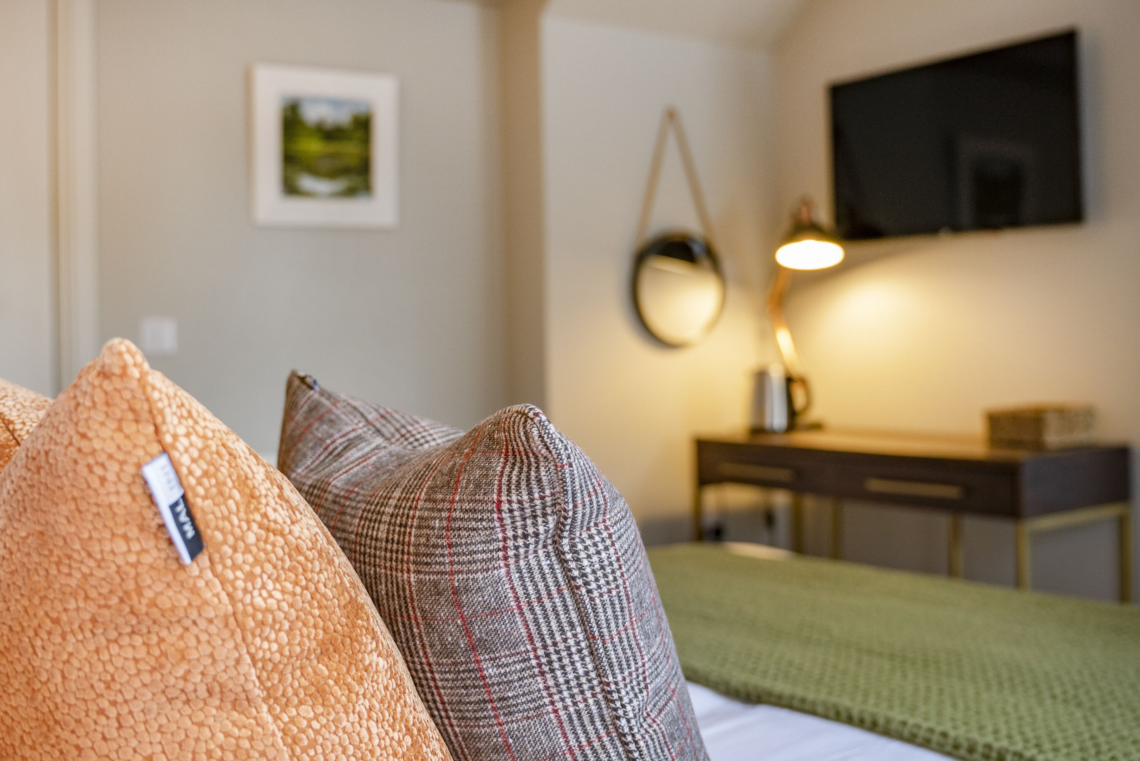 A hotel room with a bed, pillows, a desk, a lamp, and a television on the wall.