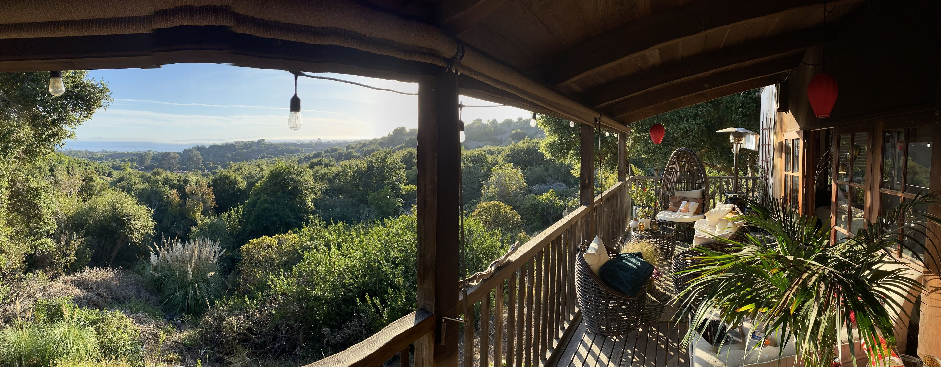 Een houten balkon met rieten meubels, een hanglamp en uitzicht op een weelderig groen landschap.