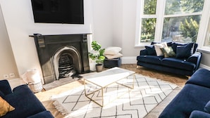 A living room with a fireplace, a blue sofa set, a coffee table, and a large window with a view of trees. - Springdale House (Buxton)