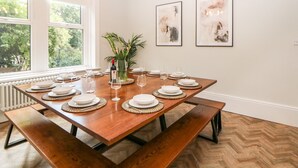 A dining room with a wooden table set for six, surrounded by benches, and a window providing a view of greenery. - Springdale House (Buxton)