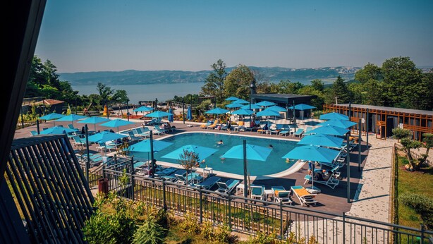 Un espace piscine doté de nombreuses chaises longues et de parasols, entouré d’une terrasse en bois et offrant une vue sur un lac et des collines lointaines.