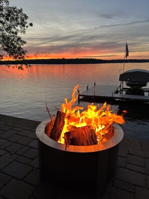 Ein Lagerfeuer mit Flammen, ein Steg und ein ruhiger See bei Sonnenuntergang.