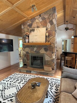 A spacious living room with a stone fireplace, a mounted television, and a dining area with wooden stools.