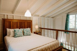 Ein Schlafzimmer mit Holz-Kopfteil, einem Bett mit gemusterten Betttextilien, einer Nachttischlampe und Blick ins Freie durch ein Fenster.