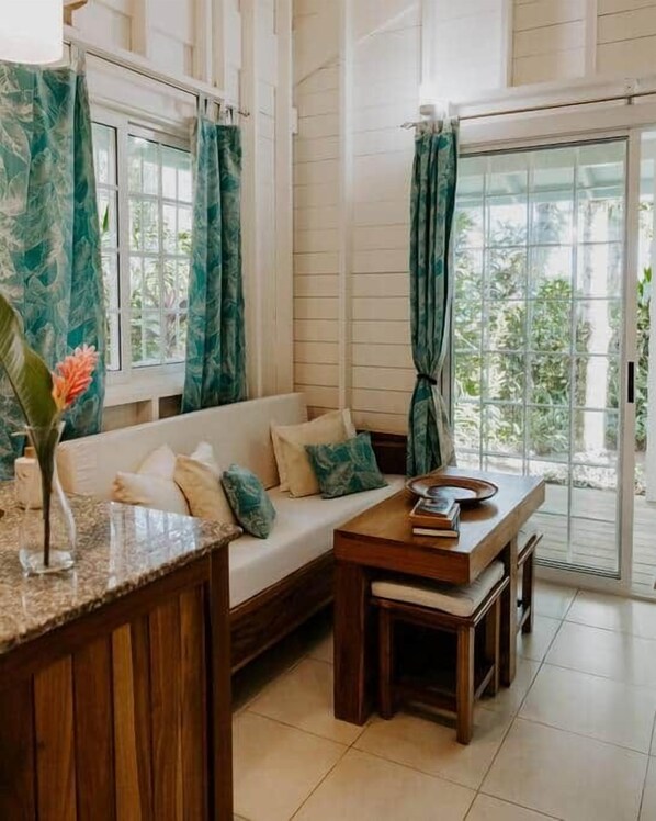 Una habitación con un banco de madera y un sofá, una mesita con un libro y un cuenco, y una ventana con cortinas.