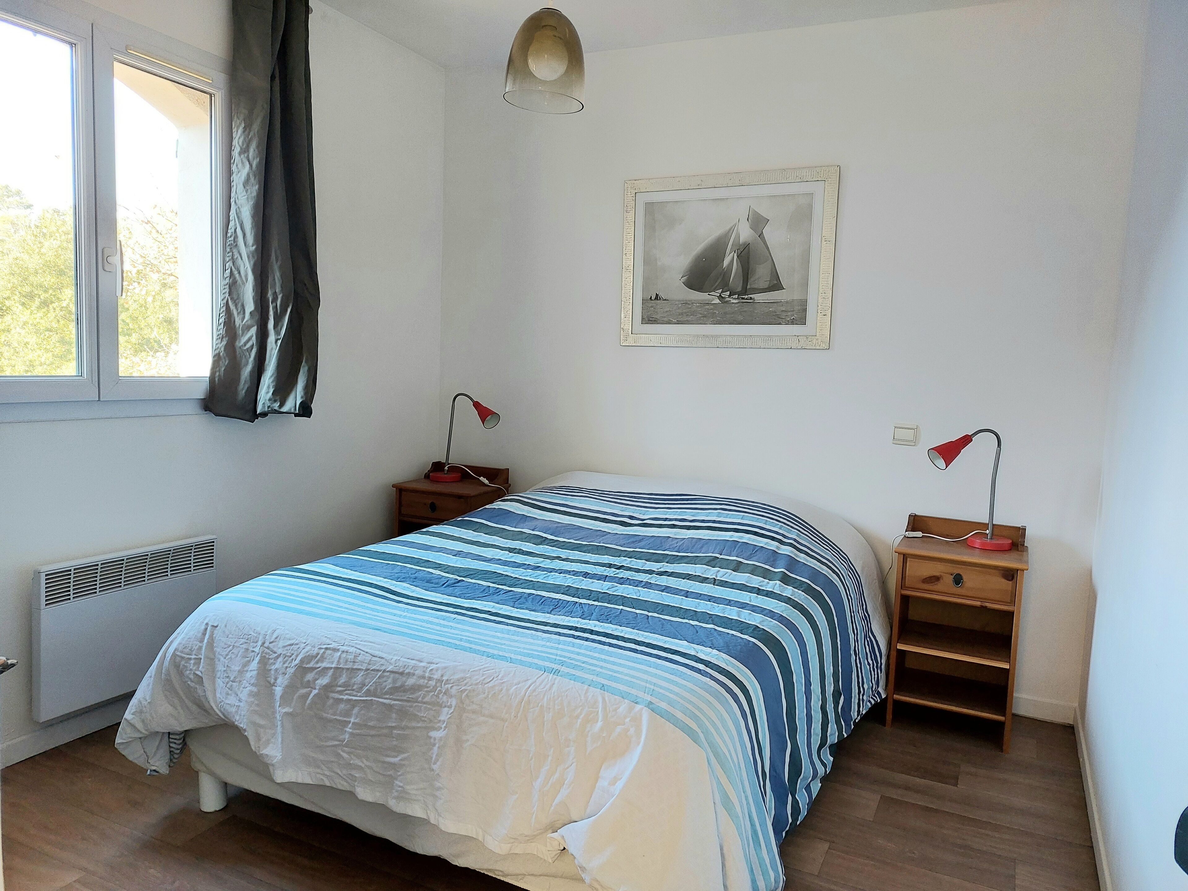 Une chambre à coucher comprenant un lit, deux tables de chevet, un radiateur, une fenêtre avec des rideaux et un tableau encadré accroché au mur.