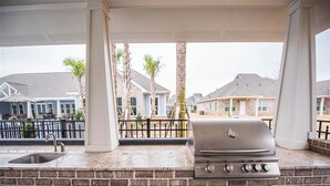 An outdoor kitchen with a grill, sink, and storage cabinets, overlooking a residential area with houses and palm trees. - Comfy Cottage (Mobile)