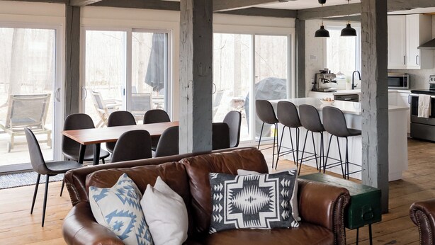 Un vaste espace de vie comprenant un canapé en cuir marron, une table basse en bois et une table à manger avec des chaises. On aperçoit au fond une cuisine avec des meubles blancs et un grand îlot central.