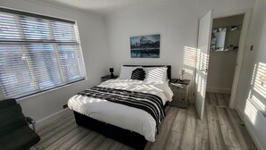A bedroom with a bed, a nightstand, a lamp, a painting on the wall, and a window with blinds. - Top Luxury 2 bed Apartment - London (London)