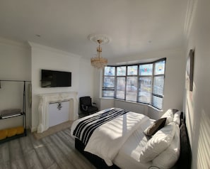 A bedroom with a large bed, a fireplace, a TV, and a bay window with blinds.
