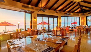A restaurant with multiple tables set for dining, large windows offering ocean views, and ceiling fans.