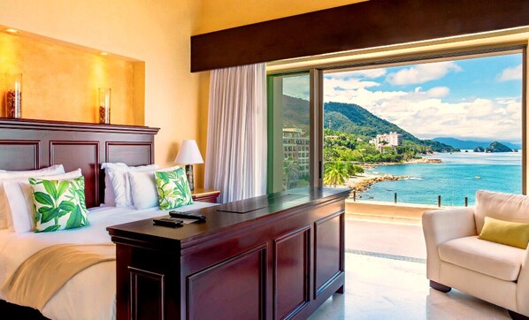 A hotel room with a bed, a desk, a chair, and a large window offering a view of a coastal landscape.