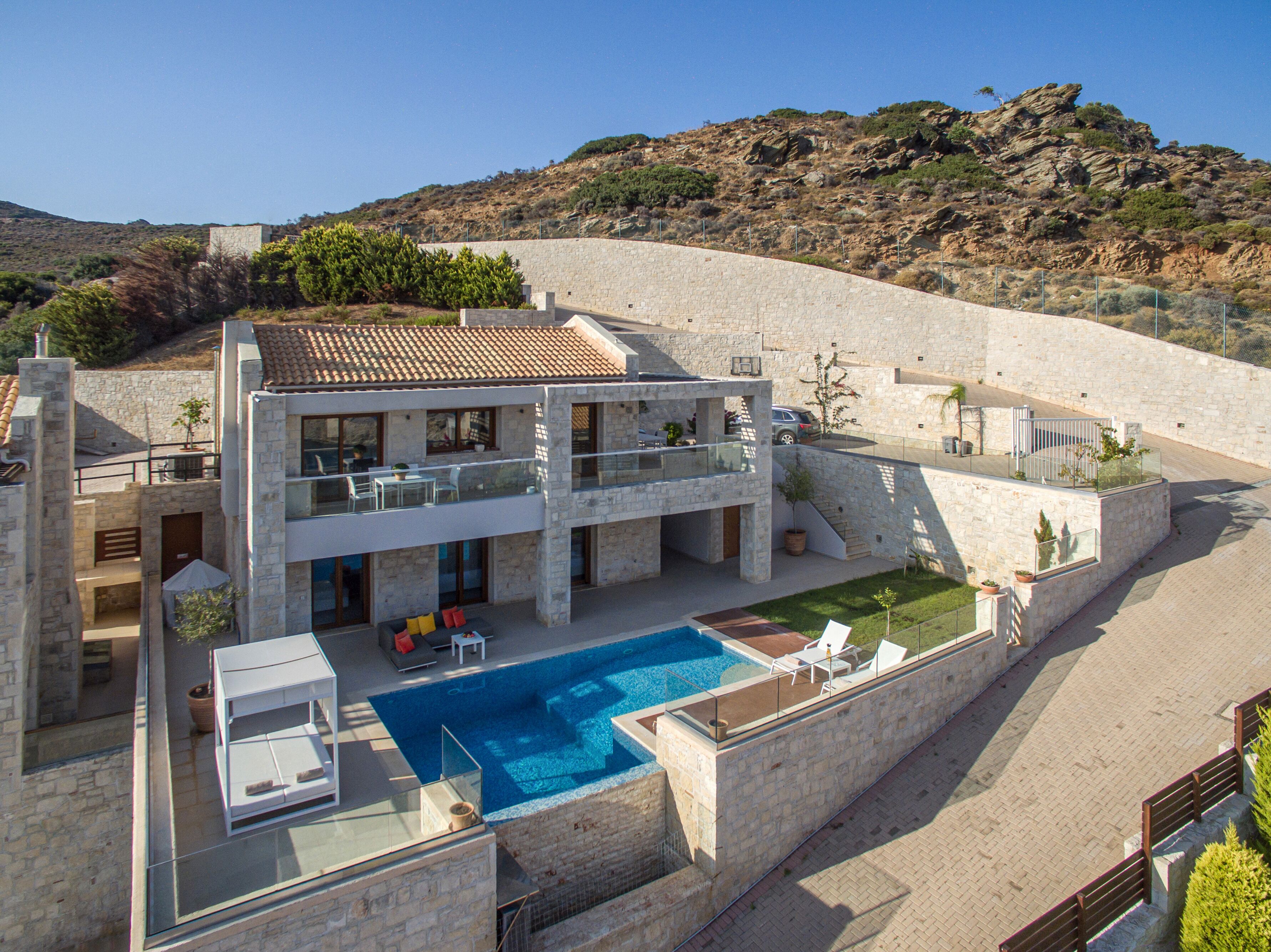 A modern two-story house with a swimming pool, stone walls, and a view of a hilly landscape.