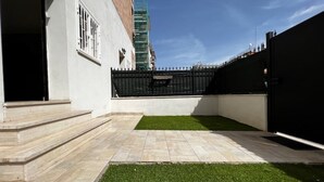 Un patio con suelo empedrado, una pequeña sección de césped artificial y una cerca de metal negro.