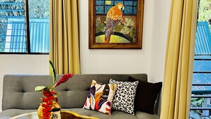 Uma sala de estar moderna com um sofá cinzento, almofadas coloridas, uma mesa de centro de madeira com uma planta e um quadro de um papagaio na parede.