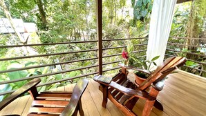 Um deck de madeira com duas cadeiras de descanso e uma pequena mesa, com vista para um jardim exuberante com diversas plantas e árvores.