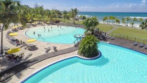 Uma piscina grande e sinuosa com uma ilha central, rodeada por palmeiras e guarda-sóis de praia.