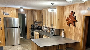 A kitchen with wooden cabinets, a stainless steel refrigerator, a coffee maker, and a clock on the wall. - Charming Fall Cabin on Lake Mille Lacs; Relax, Fish & Watch the Sunset (Isle)