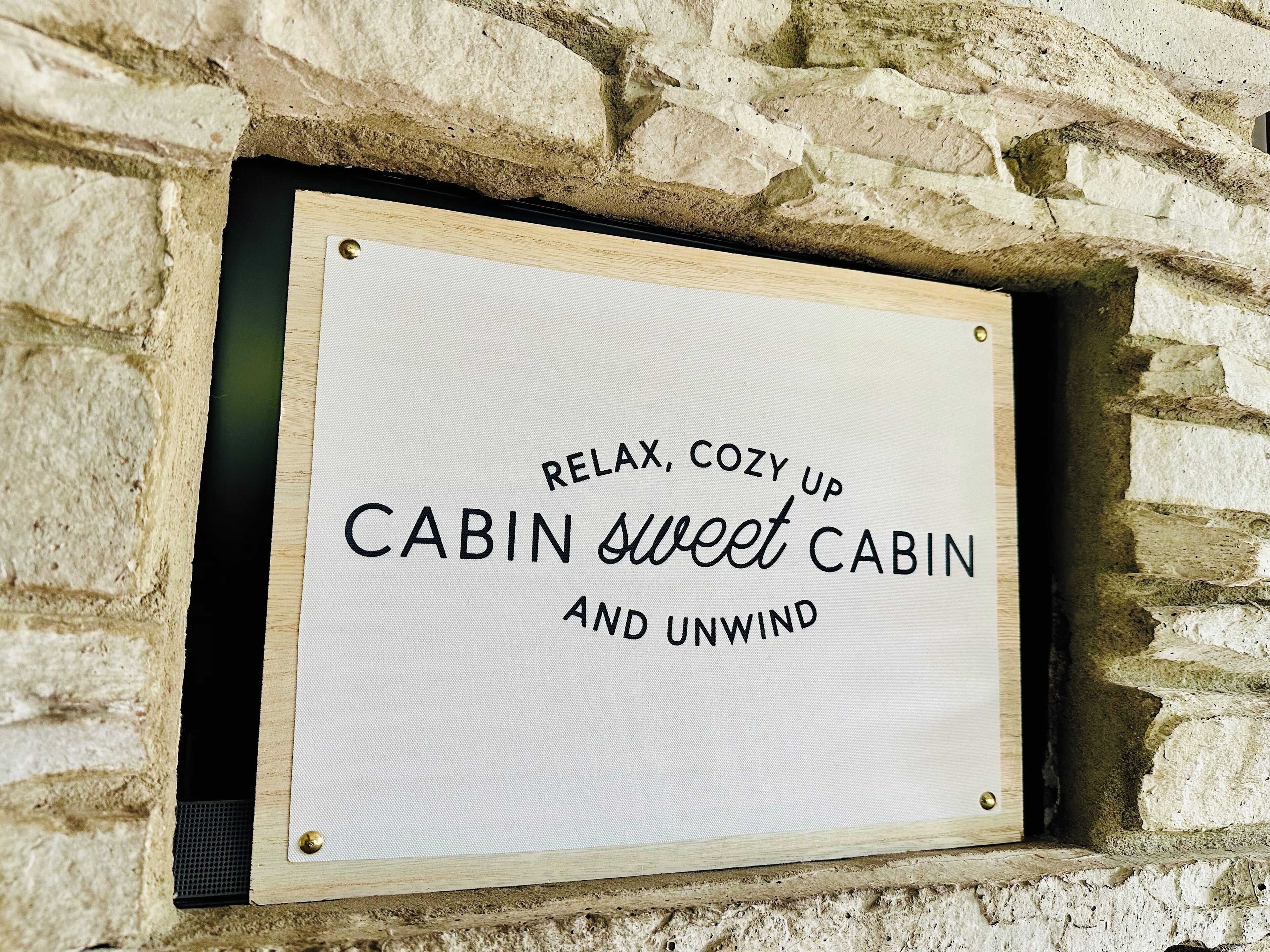 A sign with the text "RELAX, COZY UP CABIN sweet CABIN AND UNWIND" mounted on a stone wall.