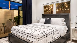 A modern bedroom with a large bed, a nightstand, a lamp, and a view of the outdoors through a window.