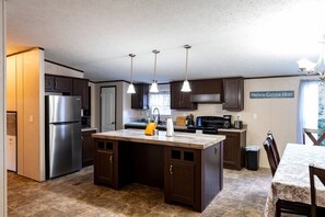 A modern kitchen with dark wood cabinets, a stainless steel refrigerator, a central island with pendant lighting, and a sign reading "Friends Gather Here."