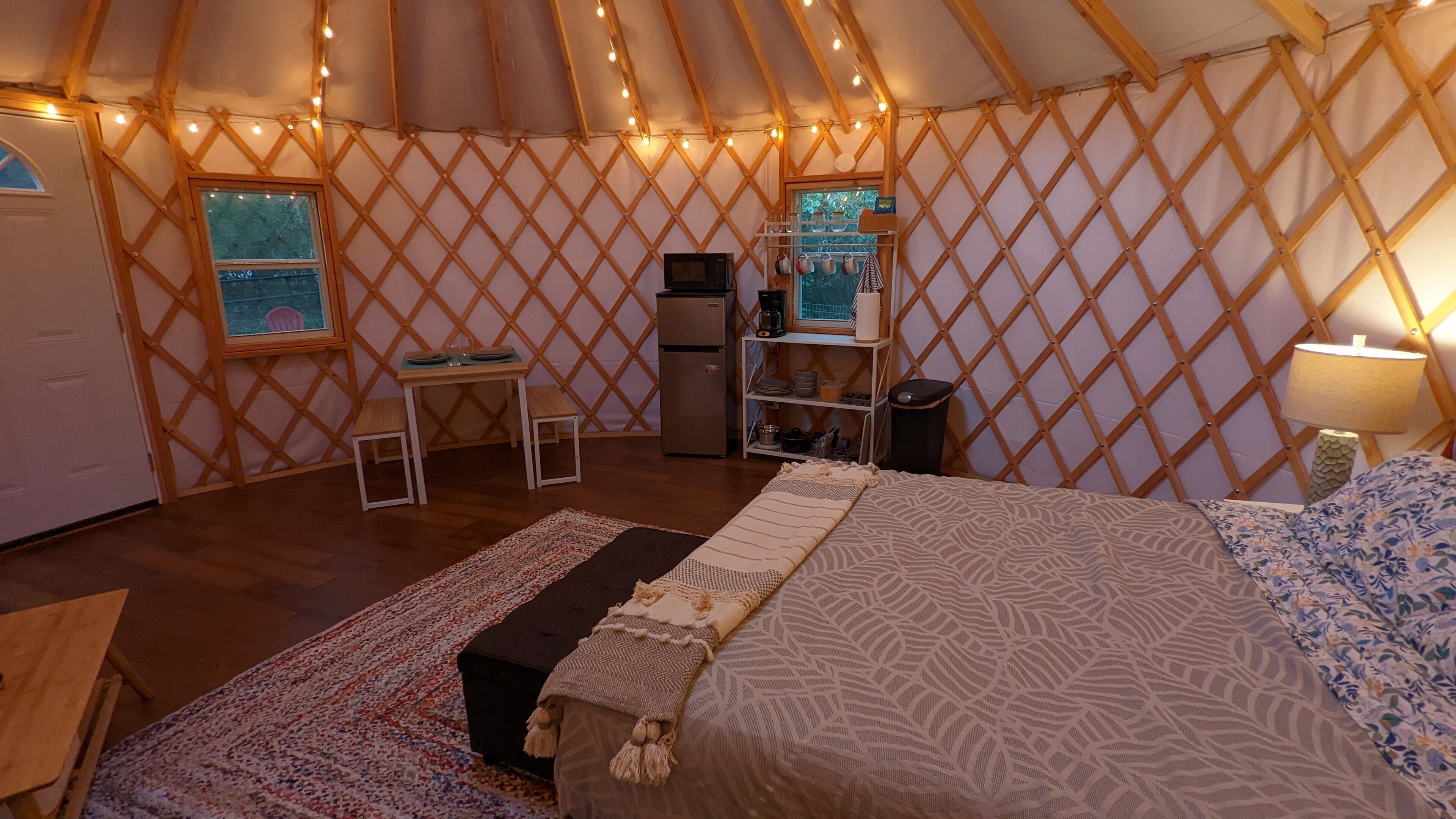 A cozy yurt with a bed, a small table, a refrigerator, and a shelf with kitchenware.