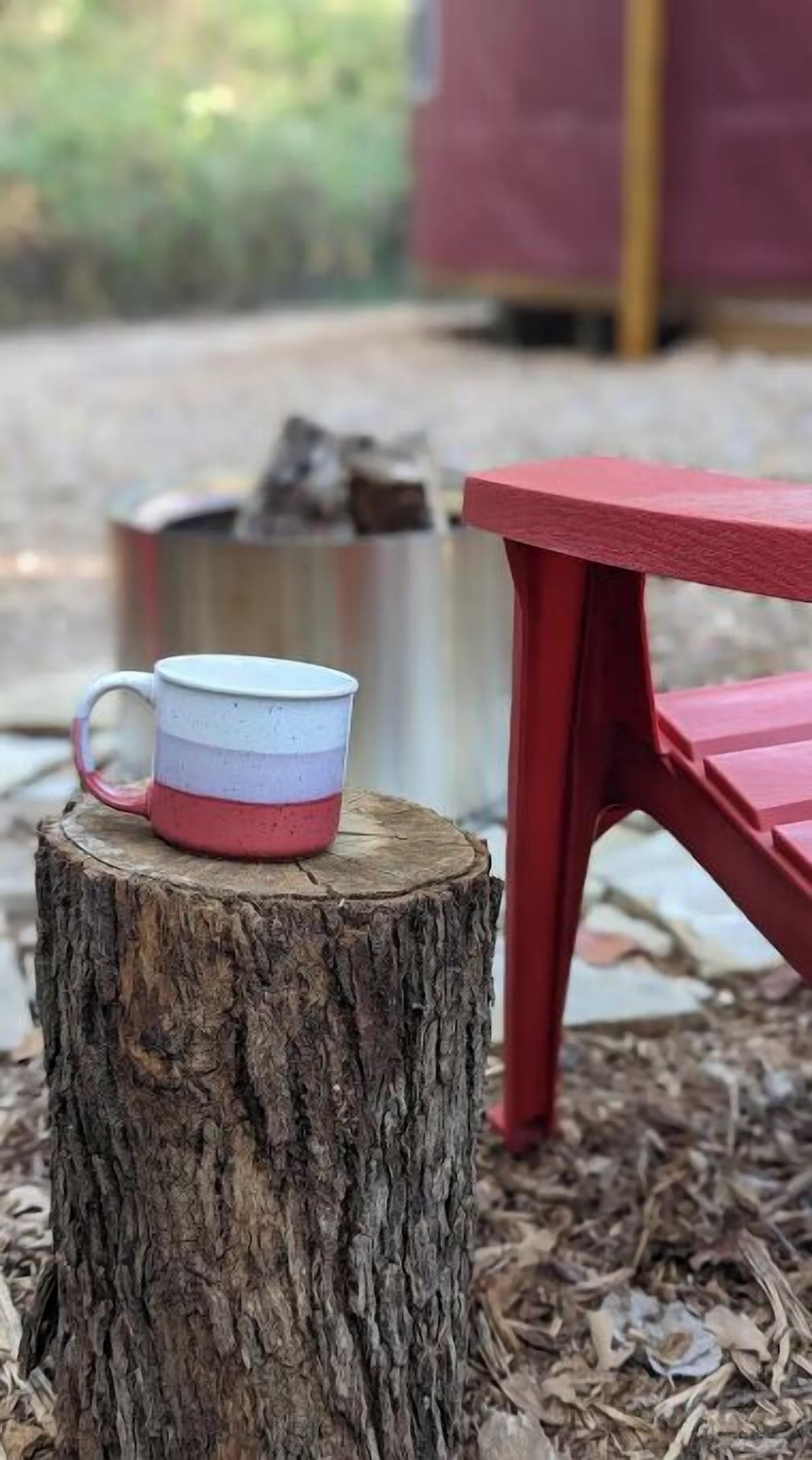 A rustic outdoor setting with a wooden stump, a red chair, and a white mug with pink and blue stripes.