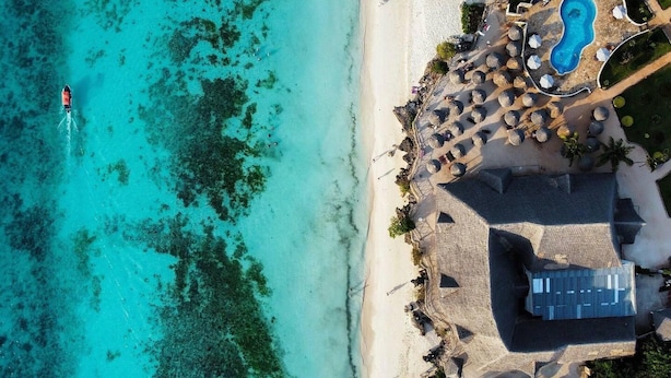 An aerial view of a beach resort with a swimming pool, beach umbrellas, and a building with a sloped roof.