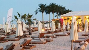 A beach resort with sun loungers and umbrellas, palm trees, and a covered area.