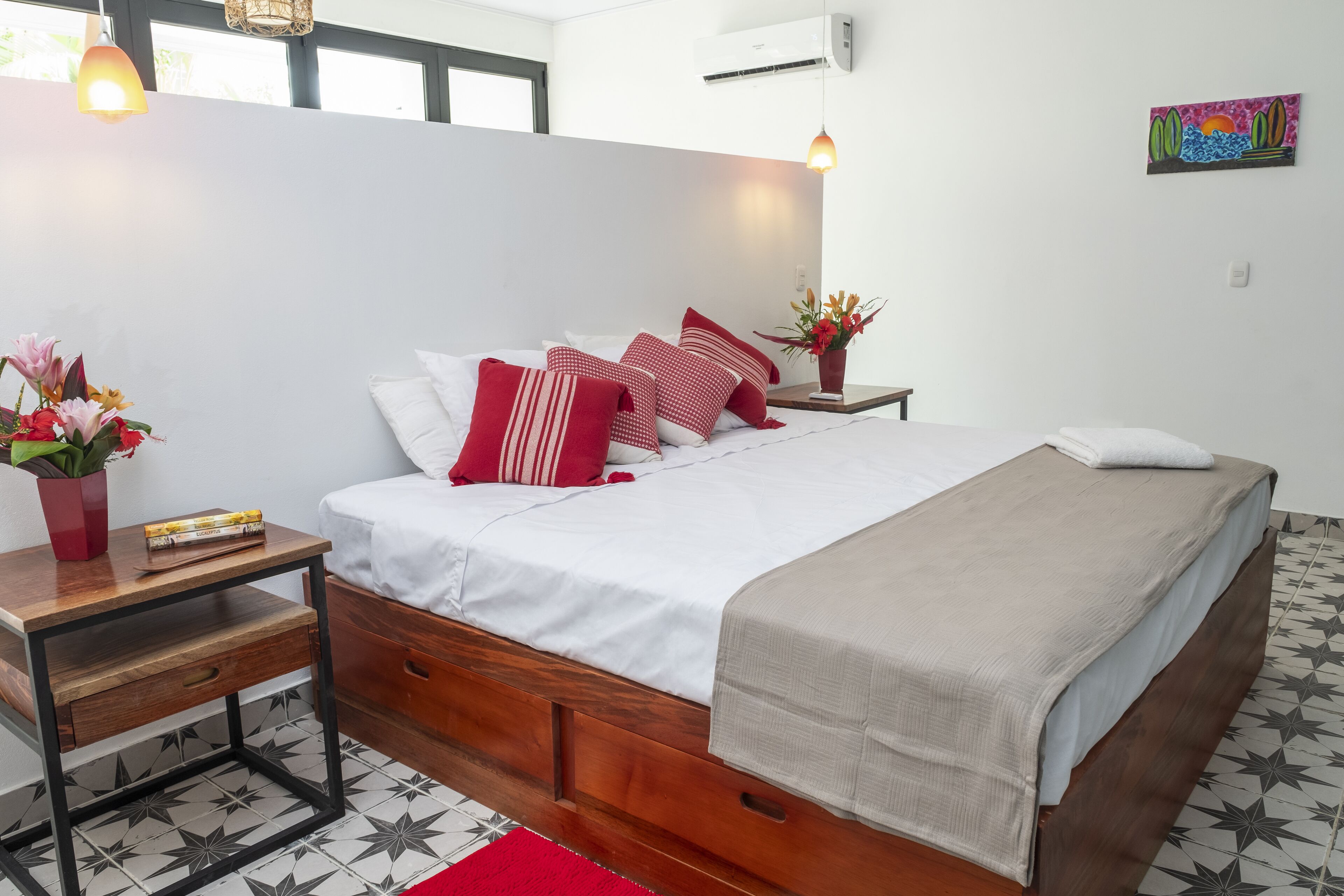 A neatly arranged bedroom with a wooden bed, white bedding, and a red rug. There is a nightstand with a lamp and a vase of flowers.