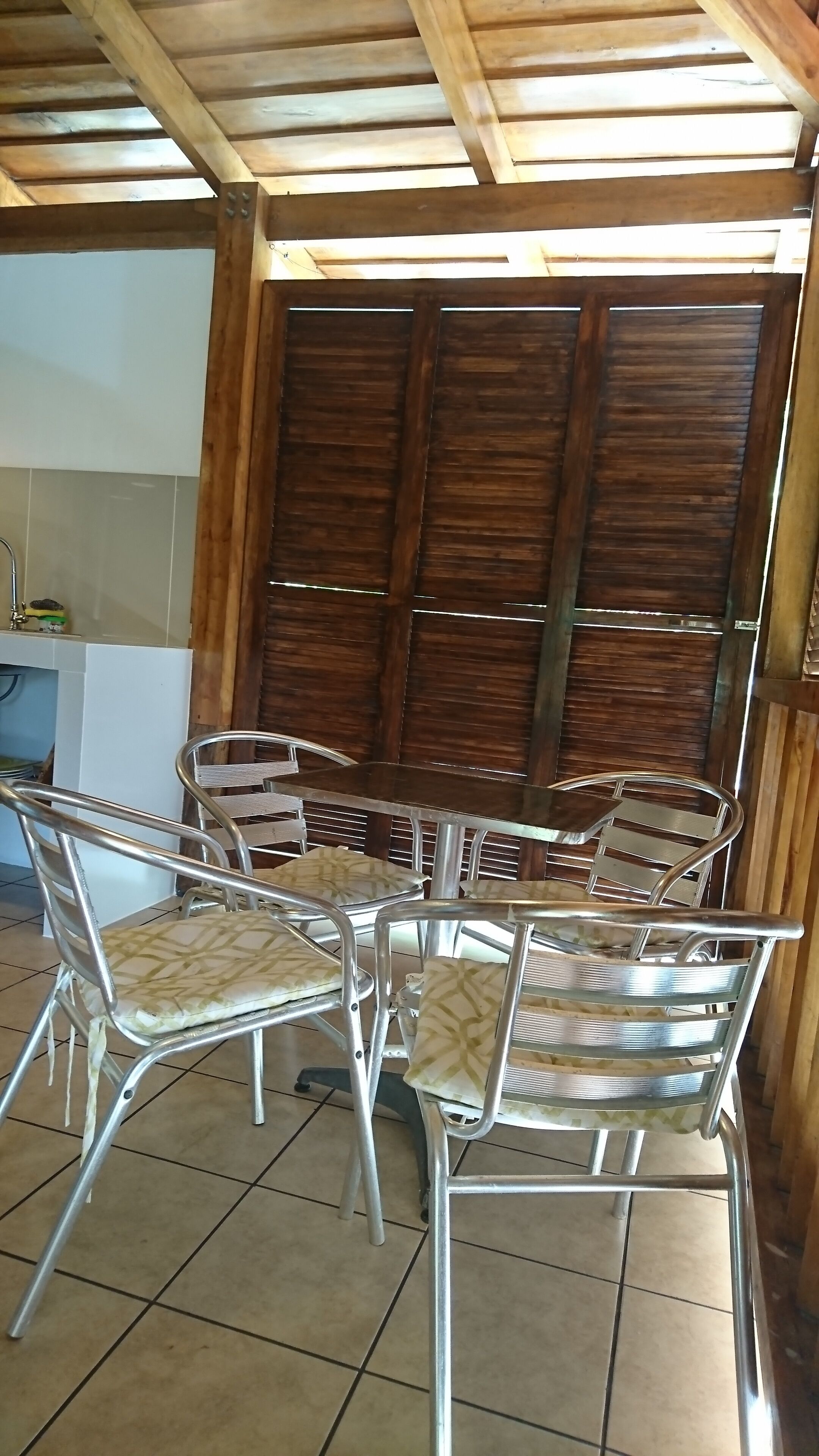 A covered outdoor dining area with metal chairs and a table, wooden ceiling beams, and a tiled floor.