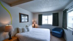 Une chambre d’hôtel avec un lit, une chaise, une table de chevet avec une lampe, une fenêtre avec des rideaux et un tableau au mur.