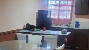 Un comedor con una mesa y sillas de vidrio, una cocina al fondo y suelo de baldosas.