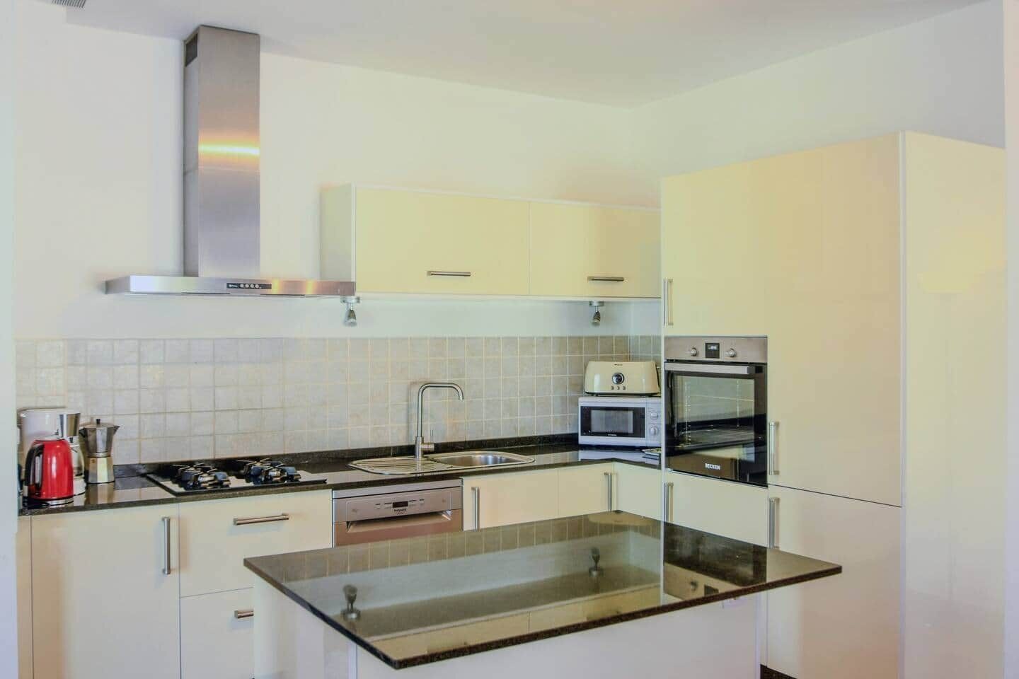 A modern kitchen with white cabinets, a stainless steel range hood, a built-in oven, and a glass countertop.