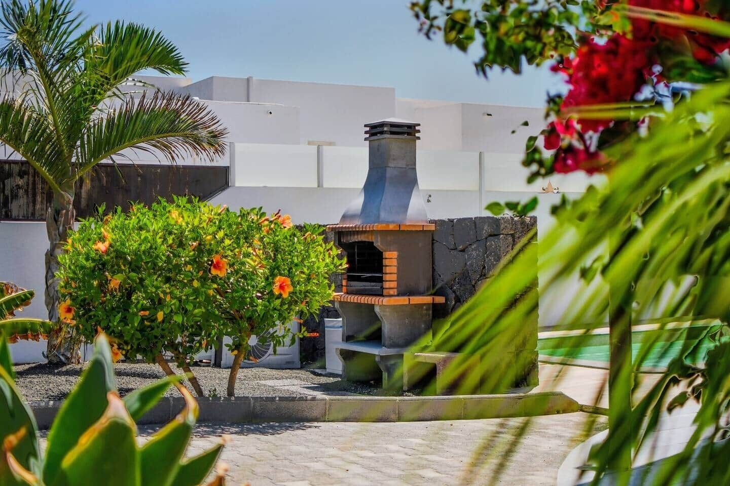 A patio area with a barbecue grill, surrounded by greenery and a white building in the background.