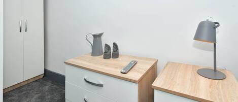 A room with a white wardrobe, a wooden desk with drawers, a grey lamp, and a remote control.