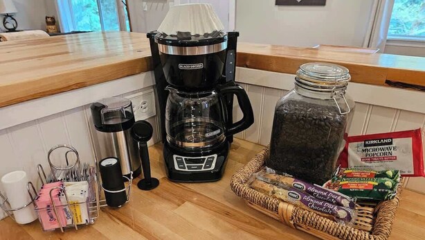 Un piano di lavoro della cucina con una macchina per il caffè, un vasetto di fagioli neri, un cestino di snack e una confezione di popcorn per microonde.
