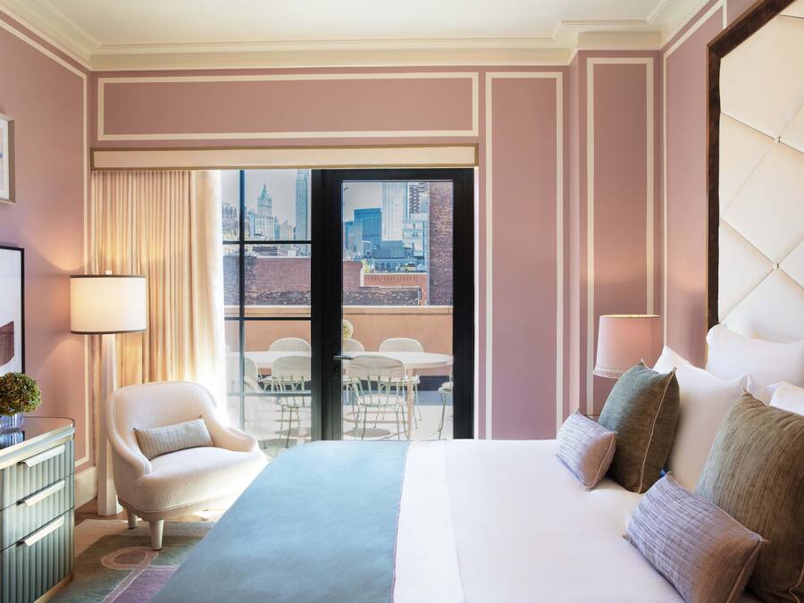 Habitación de hotel con una cama grande, una silla, un escritorio y vista a la ciudad a través de un ventanal.