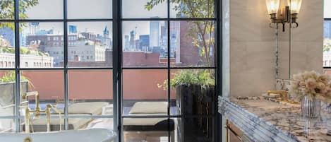 A modern bathroom with a freestanding bathtub, a large window with a city view, a marble countertop, and a wall-mounted light fixture.