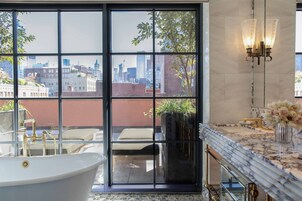 A modern bathroom with a freestanding bathtub, a large window with a city view, a marble countertop, and a wall-mounted light fixture.