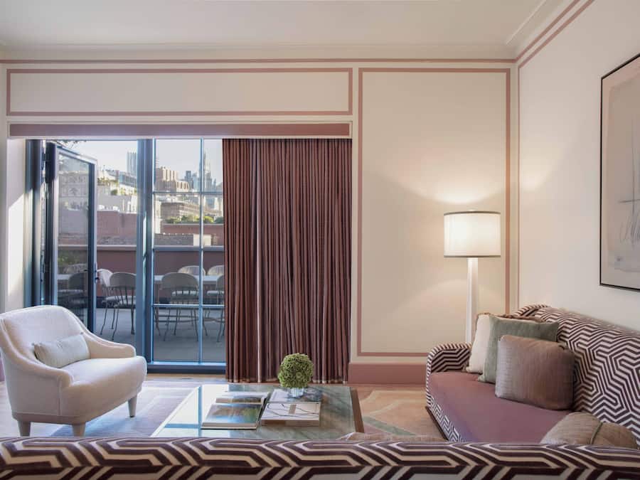 A modern living room with a sofa, armchair, and a coffee table. There is a large window with curtains, a floor lamp, and a framed artwork on the wall.