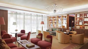 A spacious lobby with a variety of seating options, including red armchairs and wicker sofas, a central coffee table, and a chandelier.