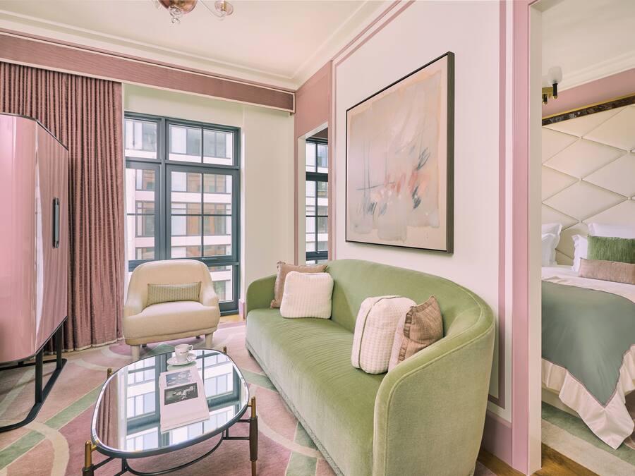 A room with a green sofa, a small table, a bed with a green bedspread, a pink armchair, and a large abstract painting on the wall.