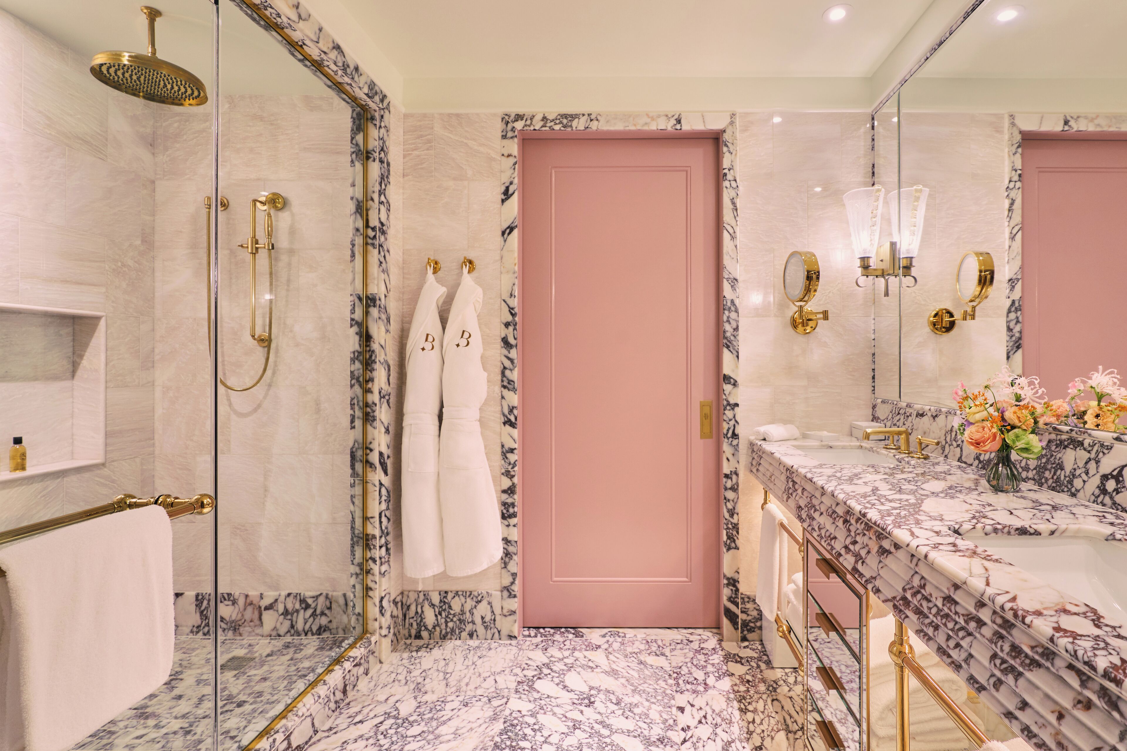 Une salle de bain avec un sol en marbre, une porte rose et une douche dotée d’accessoires dorés.