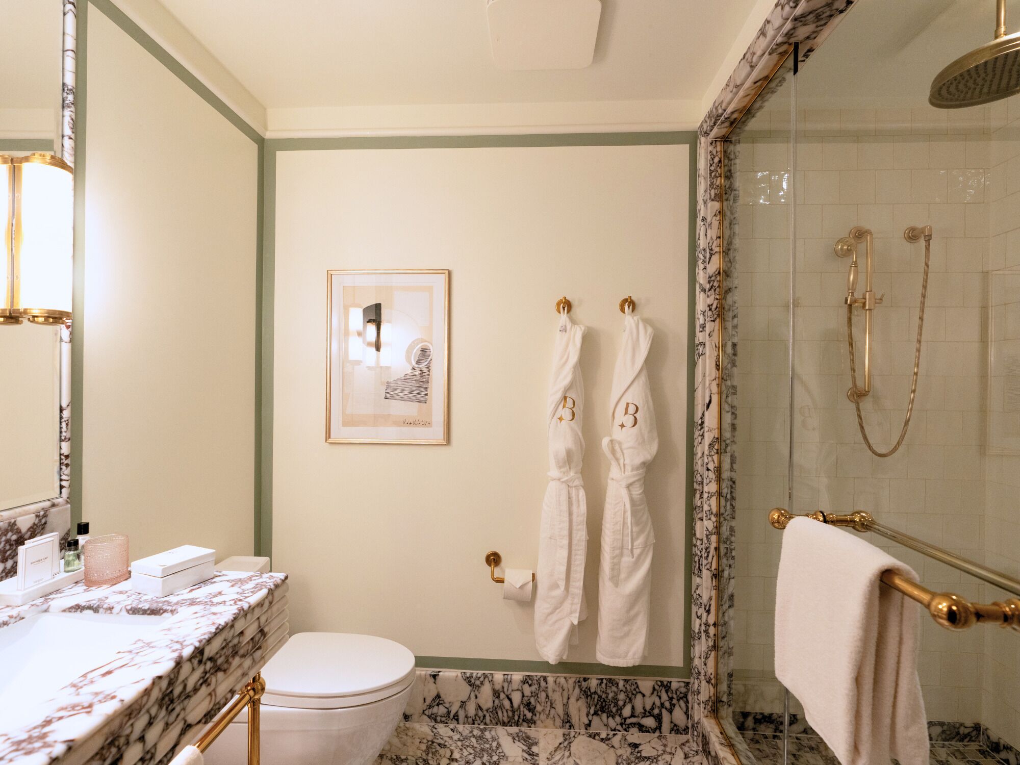 Un baño con tocador de mármol, inodoro, una obra de arte enmarcada, dos toallas y ducha con detalles dorados.